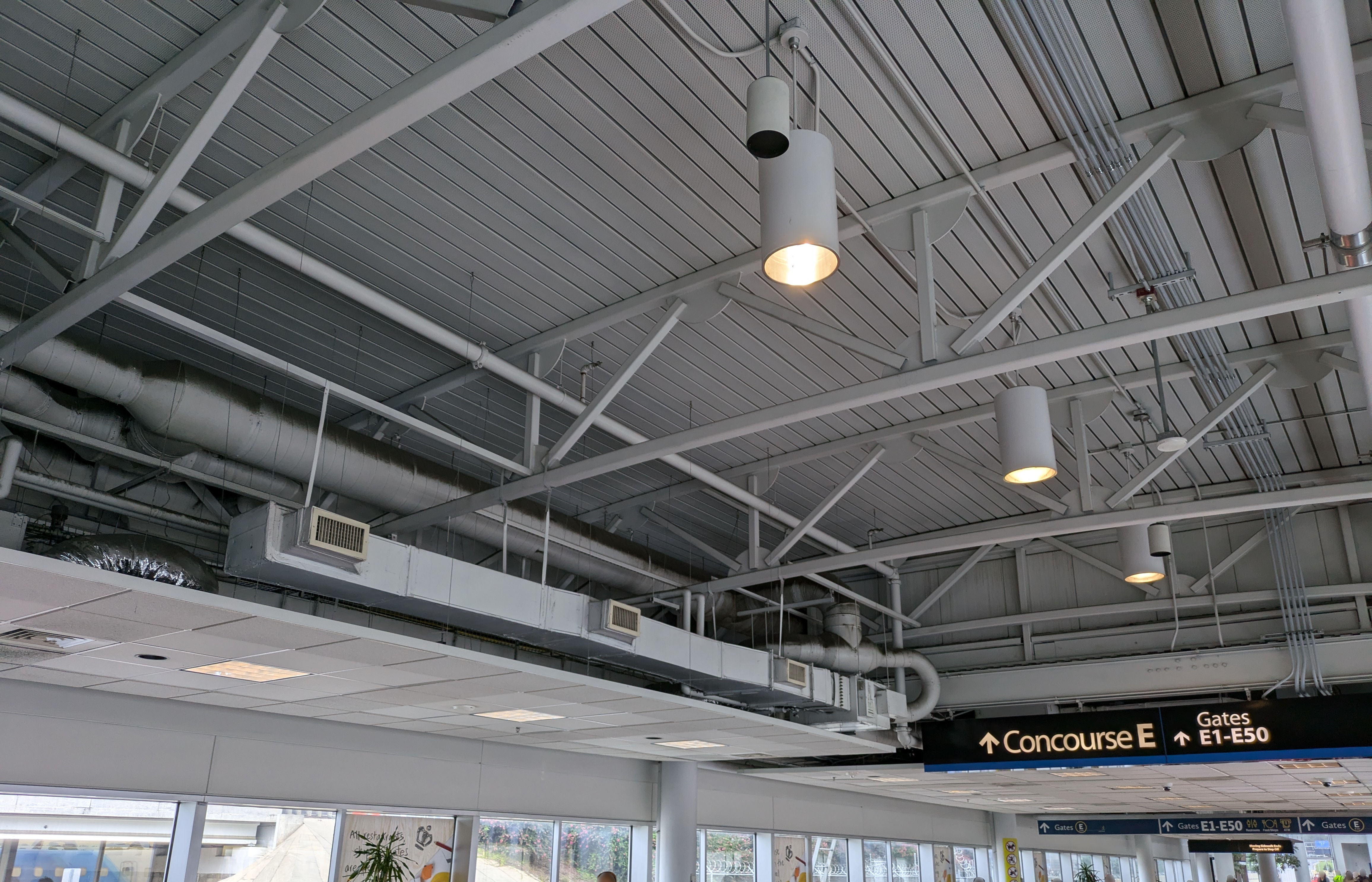 Exposed ventilation, piping, lighting, and cabling at the CLT airport looking out on a sign that pointing ahead, reading "Concourse E - Gates E1-E50".